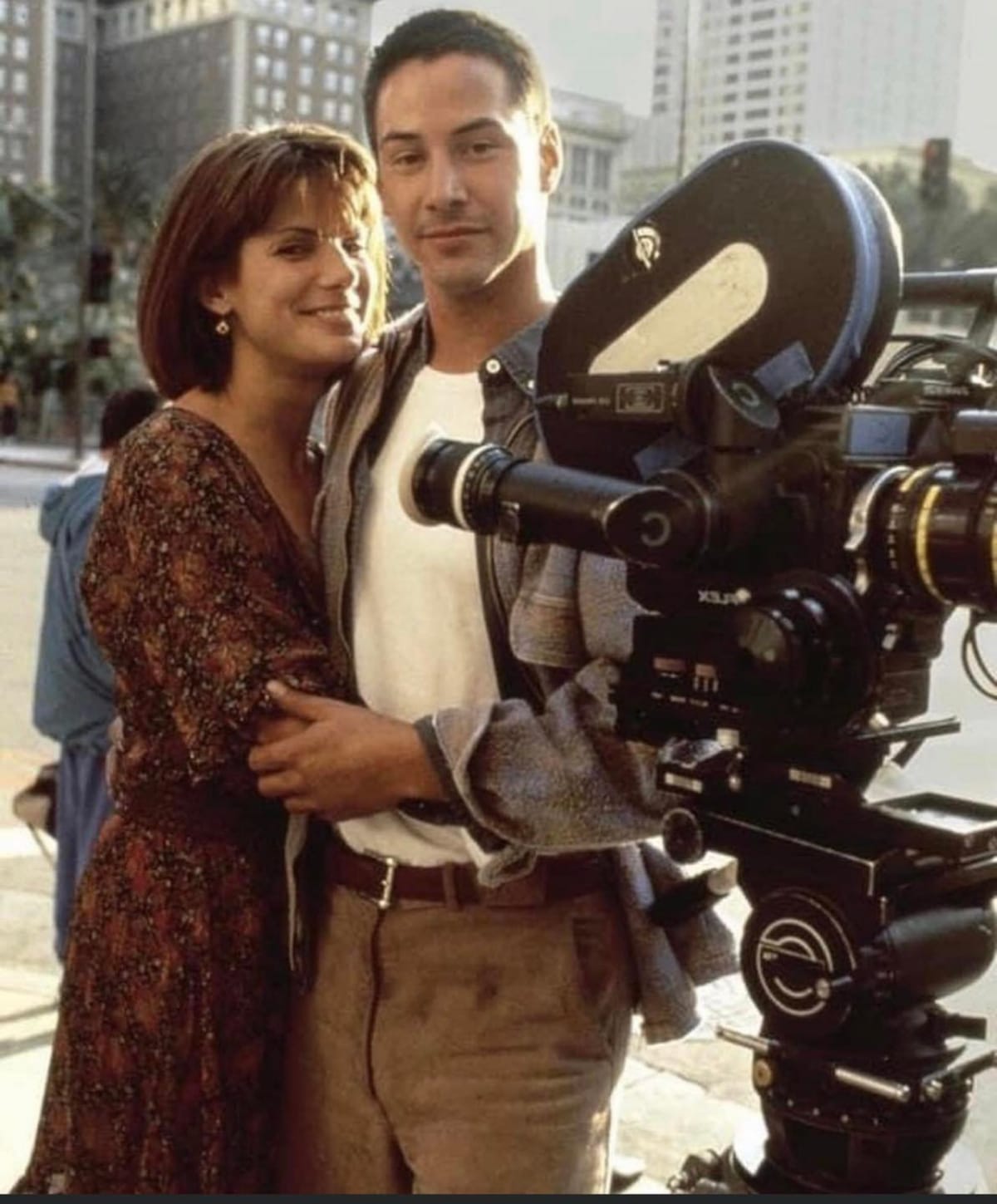 Sandra Bullock and Keanu Reeves hug and smile for the camera in their Speed costumes, standing beside a black film camera